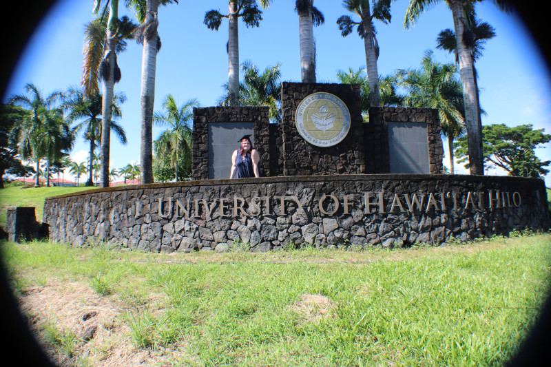 Dani Wilde standing in front of the rock UH Hilo sign leaning on the front rock wall. She is wearing a blue dress and a graduation cap and smiling.