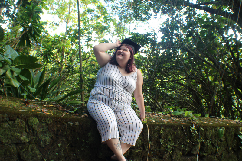 Dani Wilde sitting on a rock wall with a lot of greenery in the background. She is wearing a white striped jumper and has her right arm up holding the tip of a graduation cap on her head.