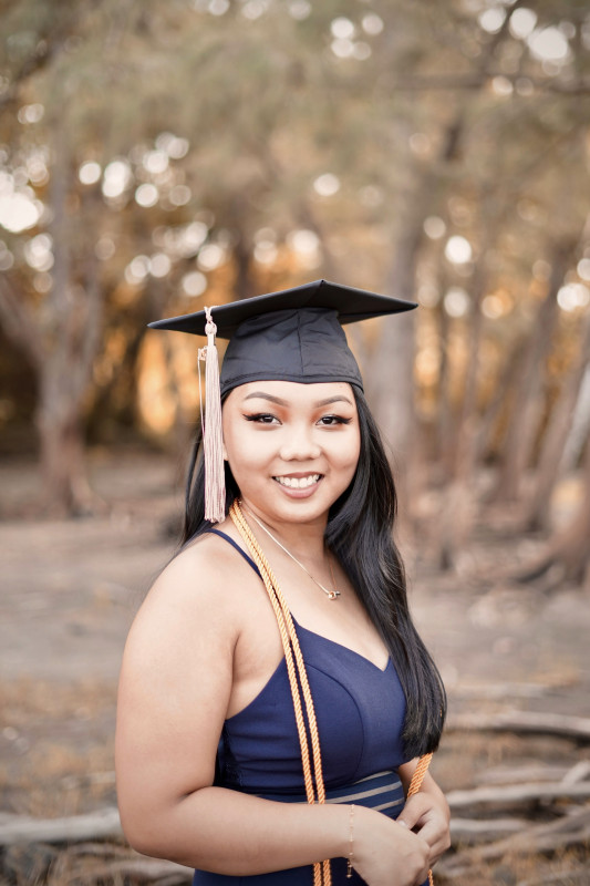 Primary photo; a half body image of me with my body slightly sideways while facing the camera. I am smiling with my teeth showing, and holding my fingers with my arm bent on 90 degrees. I am wearing a blue dress with a cap and gold cord hanging on my shoulders.