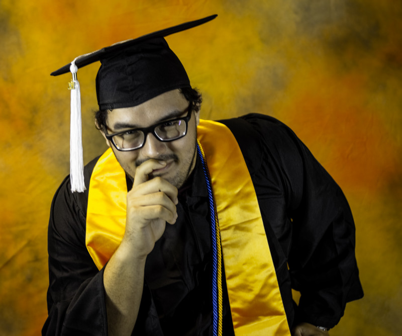 Photo is of me sitting. I am in my cap and gown with a stole and cords of Phi Theta Kappa colors (blue and gold). I am doing a pose where it is similar to a “thinker” position and you can see a smile behind my hand.