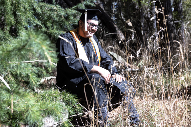 I am in my cap and gown with stole and cords. I’m sitting in a forest environment with pine trees behind me. I am smiling with enthusiasm and sun shining in my face.