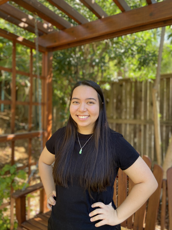 Woman in black shirt with hands on hips smiling in front of a garden.