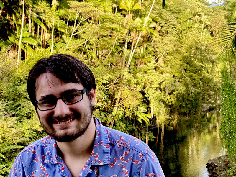 A young man wearing glasses with brown hair and a short beard wearing a blue shirt with Koi fish on it. Behind him there is a river.