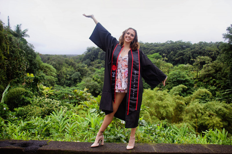 Photograph of Rachel with Akaka Falls park in the background. Rachel is in the center of the photo with her arms up wearing the graduation outfit and Marine Science custom stole.