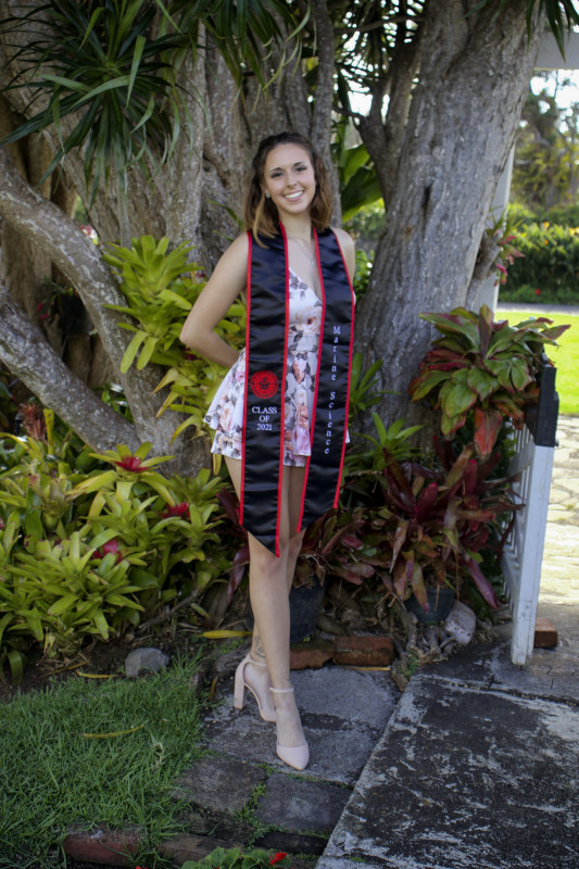 Photograph of Rachel with plants in the background. Rachel is in the center of the photo wearing a white romper and Marine Science graduation stole.