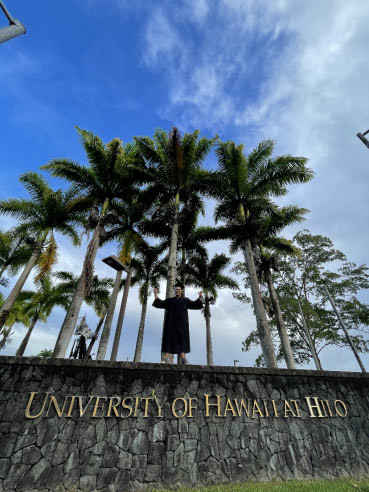 Standing on the sign in front of UH Hilo campus in regalia