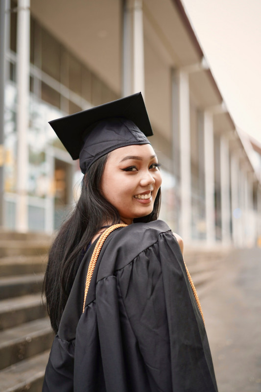 I am turned smiling towards the camera with my body facing the other way. On my shoulder are my gown and cord hanging while I hold on to them. I have my cap under my straight black to dark brown hair. In the background is the school's office building.