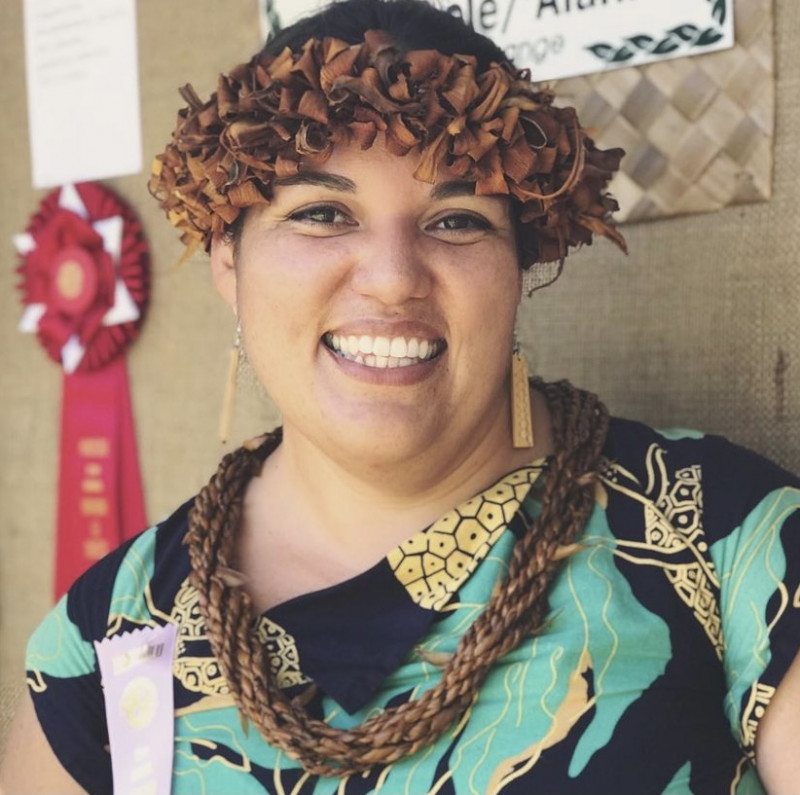 A smiling Polynesian woman with a lei po'o and colorful print dress on.
