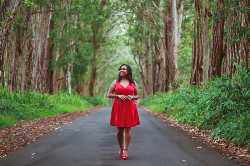 Dark shoulder length hair with dark brown skin tone. Red dress with red six inch heels. Gold chain belt, gold ring and gold chain necklace. Background is of a forest, the subject stands in the center on a paved road.