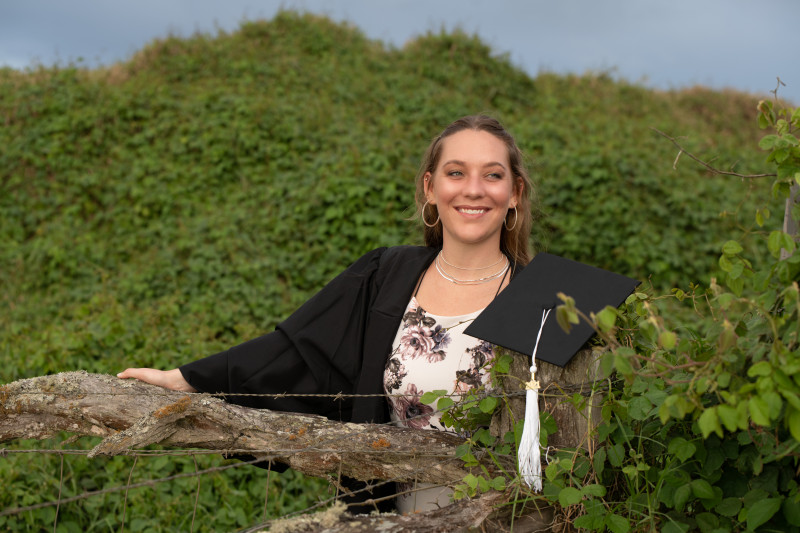 Photo of Savanna smiling and leaning on a fence post with a graduation cap hanging on the fence.