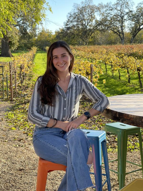 I am sitting on a colorful, metal highchair next to a round, wooden table. My hair is in front of my right shoulder, head tilted down slightly and smiling. I am wearing a grey and white striped long sleeve, collared shirt with blue jeans. Behind me, a grape vineyard separated by vibrant green grass down the rows. Behind the vineyard is an almond orchard.