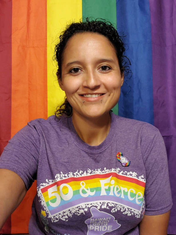 I am wearing our 2019 Hawai'i Island LGBTQ Pride shirt with the Rainbow flag hanging behind me.