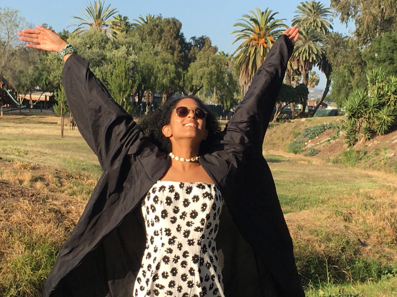 Girl wearing a black graduation gown throwing her arms up in the air for her newfound freedom.