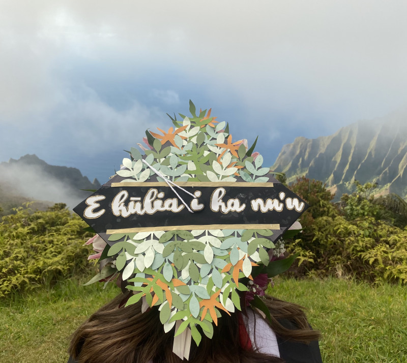 Graduate, Shantel Kalola Tamiko Rita, wearing Graduate Cap with saying, E Kulia I Ka Nu’u with the Kalalau Valley in the background