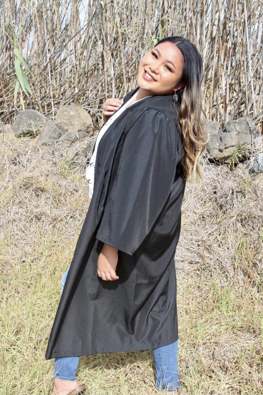 This photo was taken in a dry/dead grassy field in Waimea. I am wearing my graduation gown over a white floral long sleeve blouse, paired with blue jeans. I am smiling (with teeth) over my shoulder with my eyes barely open because of how sunny it was!