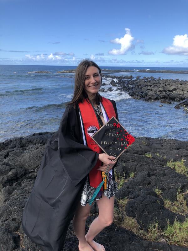 Smiling at the beach in cap, gown, stoles, and cords.