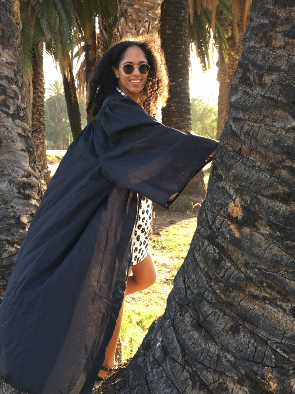 A girl named Natasha with flowing curly hair, transition glasses, wearing a white dress with black flowers. She is also wearing a black graduation gown. She is also standing in a tree