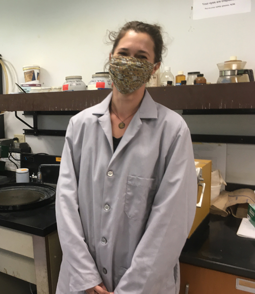 Alice is pictured wearing a grey lab coat and her face mask while standing in the geology lab. It's covered up but she is smiling under her mask.