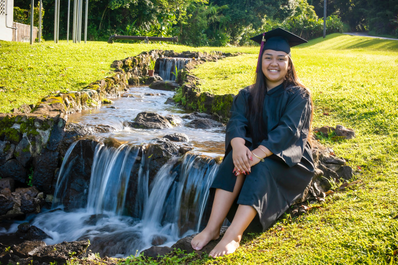 Full body, cap and gown photo taken next to a flowing waterfall.