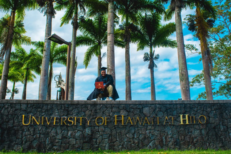 standing on top of the UH Hilo sign in full graduation attire