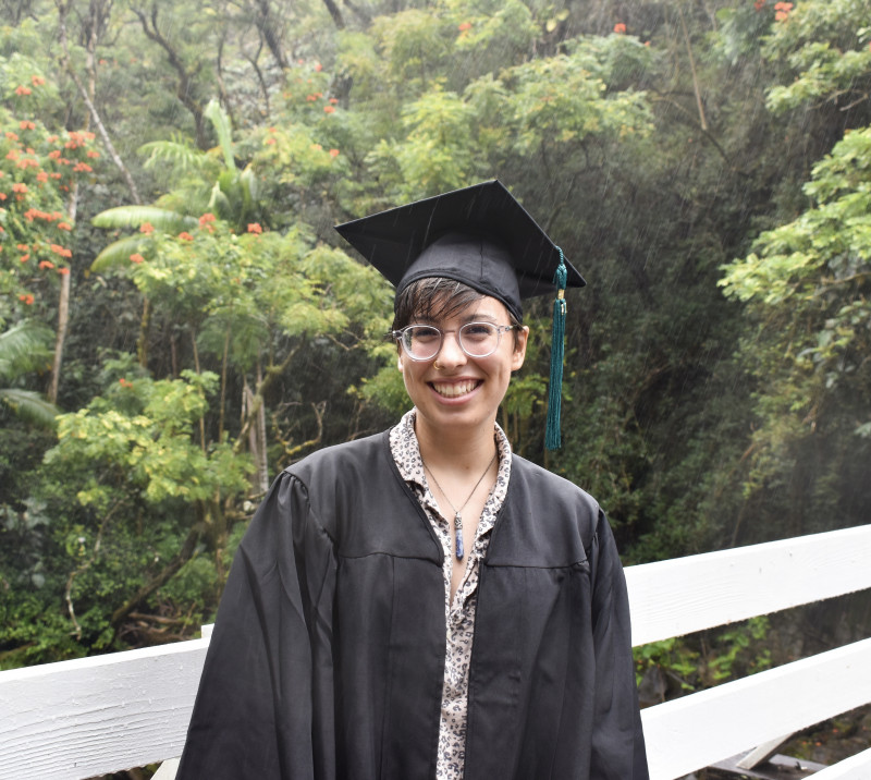 Lindsey stands on a bridge with the jungle in the background smiling at the camera in her cap and gown