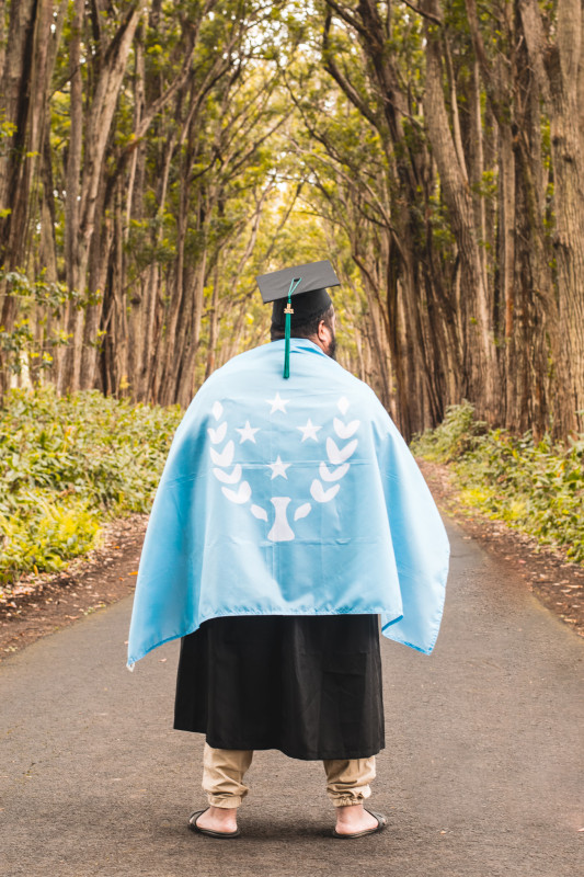 Standing amidst tall trees dressed in cap and gown with the Kosrae flag