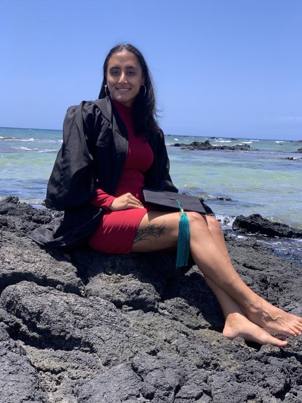 Me, wearing a red dress with my black gown on and my cap on my lap, siting down on a rock, smiling at the camera as the waves roll in behind me.