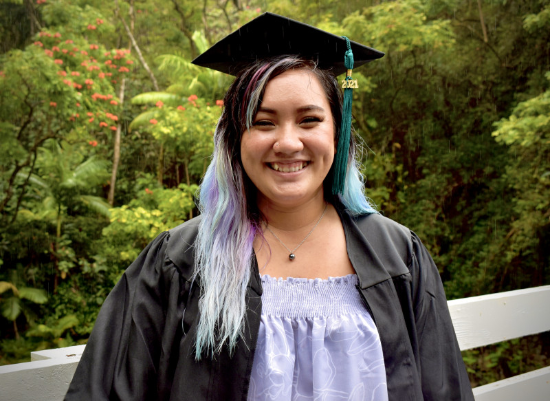 Graduate in their cap and gown in front of greenery with a huge smile.