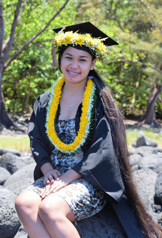 A young graduate sitting in graduation attire smiling at the camera wearing a yellow Yapese neck lei and lei haku.