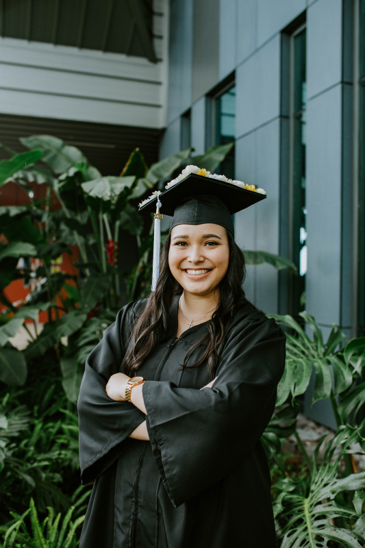 This image is of myself crossing my arms in my cap and gown with greenery (monstera leaves) in the background.