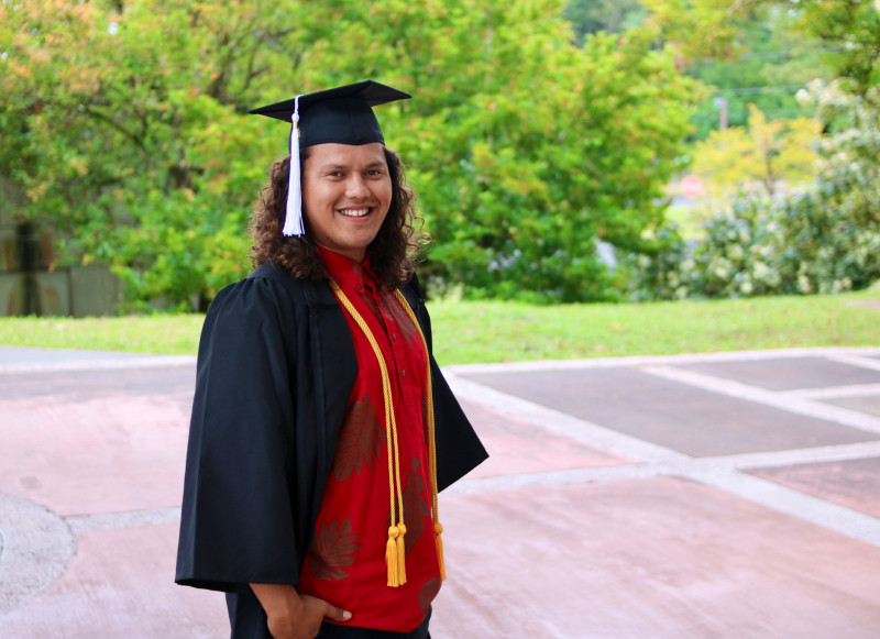 My fifth and final photo is of me with my cap and gown standing outside with my hands in my pocket