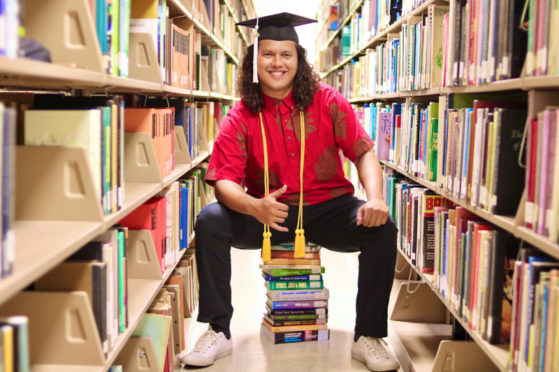 My PRIMARY PHOTO is of me sitting on a stack of books in the library in between rows with a shaka. I am wearing a red aloha shirt with black pants and white shoes.