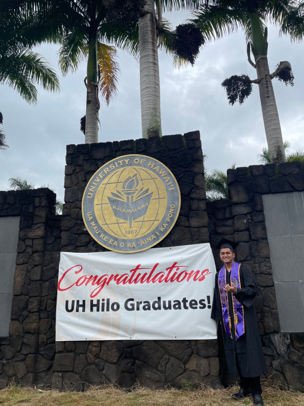 I am standing in front of the UHH rock wall sign with my black cap and gown, smiling and throwing a Shaka. Over my gown, I am wearing a purple and yellow stole and chords.