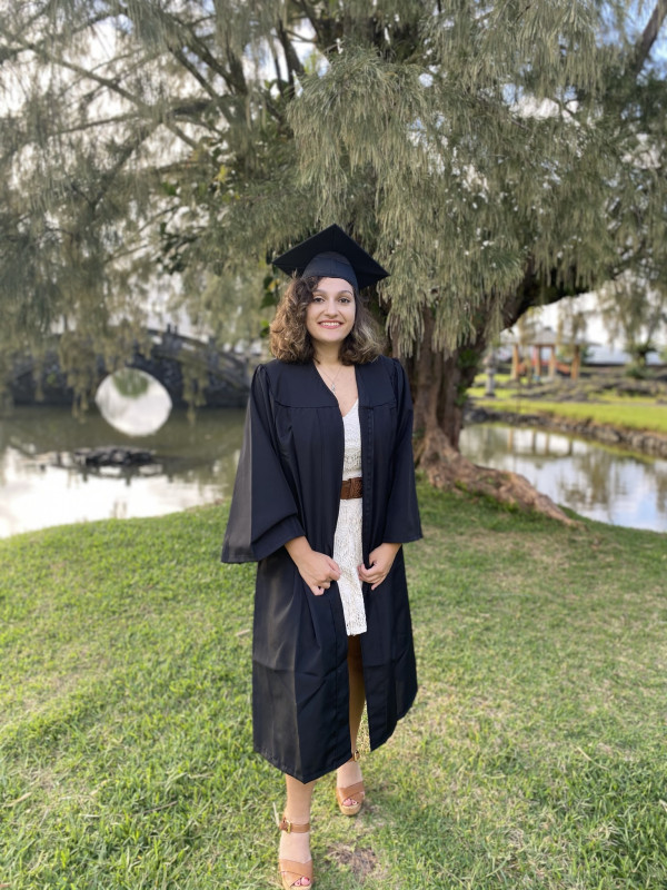 Cara is standing in a white dress and black graduation gown unzipped, graduation hat and turquoise tassel atop her head and tan heels. She is smiling. In the background is a large tree which has thin and long drooping leaves. There are two ponds and a small black bridge crossing one in the background as well.