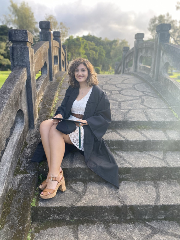 Cara sits on the steps of a small dark stone bridge. She's smiling in a white dress, the black graduation gown unzipped, tan heels, with the flat graduation hat and tassel across her lab.