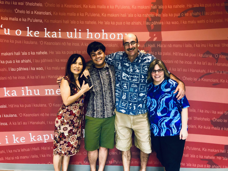 I am pictured with my arms wrapped around my three linguistics professors, Dr. Ohara, Dr. Saft and Dr. Perez. We are standing in front of a dark red mural inside of Hale ʻŌlelo which depicts Hawaiian writing.