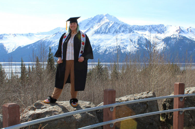 I am standing on a rock with mountains behind me. I am wearing my cap and gown with Beaded Native Regalia sole on.