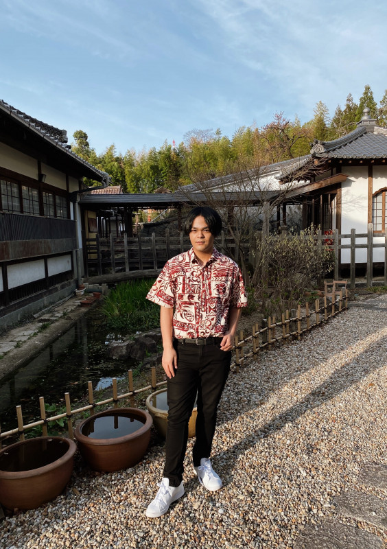 This serves as my primary photo, depicting me standing in a garden at a temple located next to my host university in Nagoya, Japan. I am wearing a white and red aloha shirt with traditional Hawaiian patterns, honu, pineapples, and flower motifs. I'm wearing black jeans with a belt as well as white Adidas shoes.