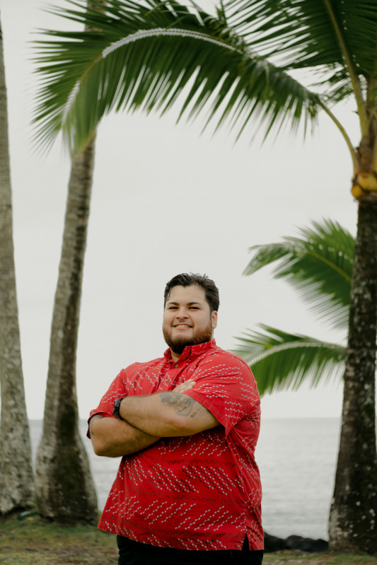 A picture of me down at Puhi Bay standing cross-arm and smiling with the ocean and palm trees as my background.
