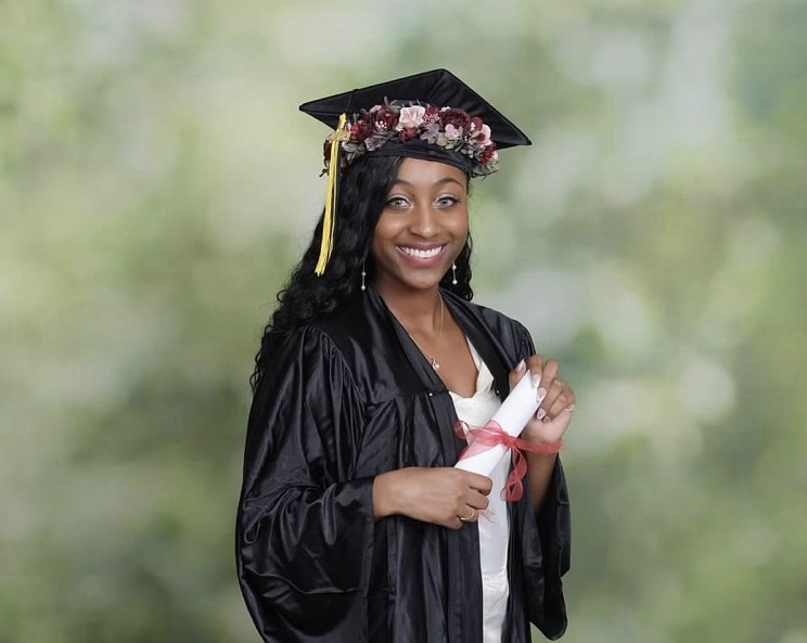 This is my primary photo that will be listed with my name and degrees earned. A picture of me smiling turned slightly to the side facing the camera. In the picture I'm wearing a white dress, black cap, black grown, and burgundy and pink flower crown with a black and gold tassel. The background of the picture is an outdoor green backdrop