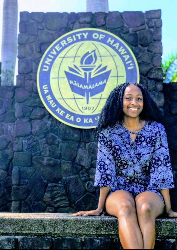Casual photo of me sitting in front of the UH Hilo logo on campus.