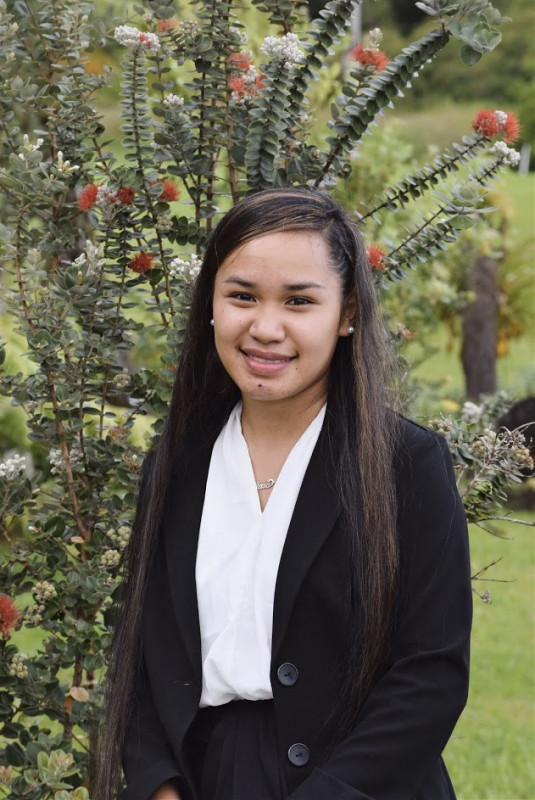 A young graduate in business attire smiling at the camera.  She is sitting in front of a red 'ohia.