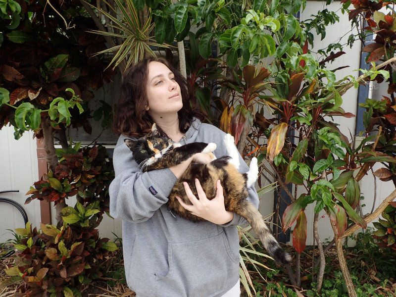 A young man with pale skin and dark hair looks into the distance, a slight smile on his face. In his arms, a chubby calico cat is bundled up; she is looking directly at the camera. In the background, a variety of plants can be seen.