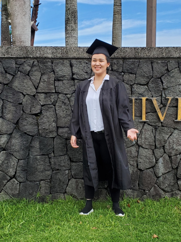 Posing and wearing my graduation cap and gown by the UH Hilo rock wall by the flag poles