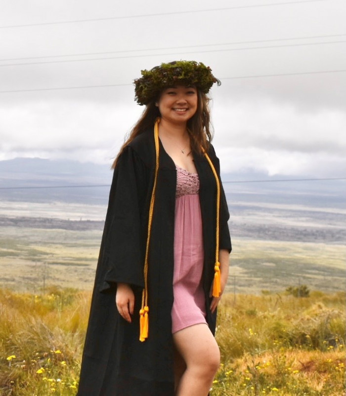 I am in a green haku, black gown, a gold cord around my neck, and a mauve dress under my gown. The background is of grassy hills with blue cloudy skies.