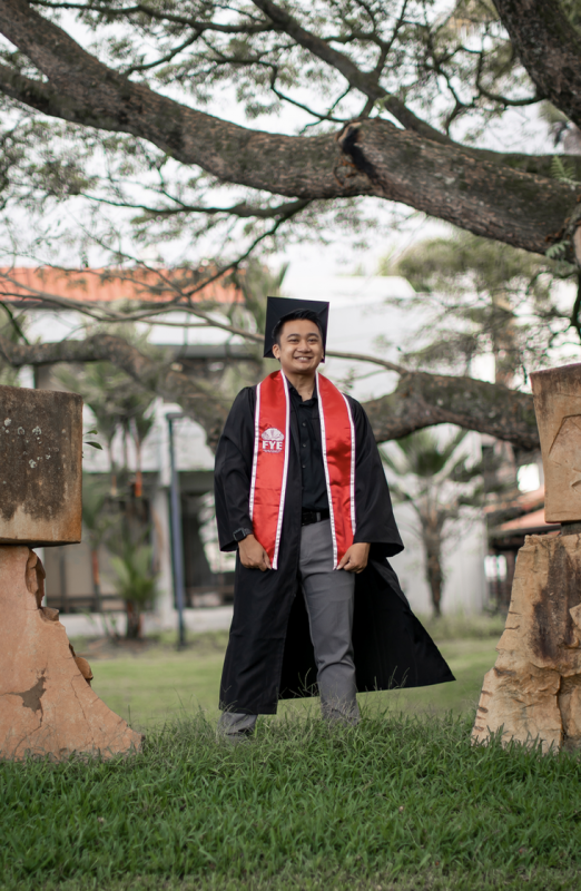 Picture of male in cap and gown in between trees on campus.