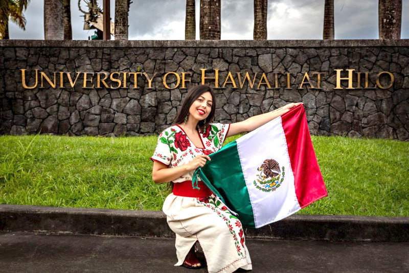 Geneba Revuelta holding a Mexican flag in front of the UH Hilo sign