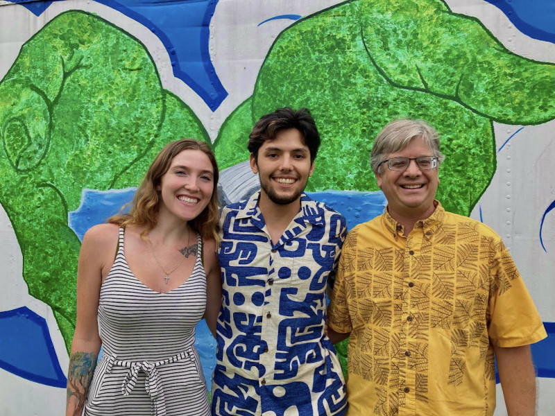Darien smiling alongside her coworker Spencer and her professor Dr. Colbert. Behind them is a painful of hands holding the ocean with a whale jumping