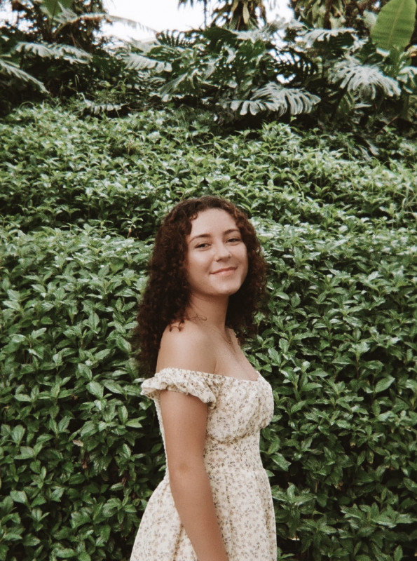 Isabella stands to the side in front of a green foliage background. She wears a yellow dress with small scattered flowers and her brown curly hair is down around her shoulders.