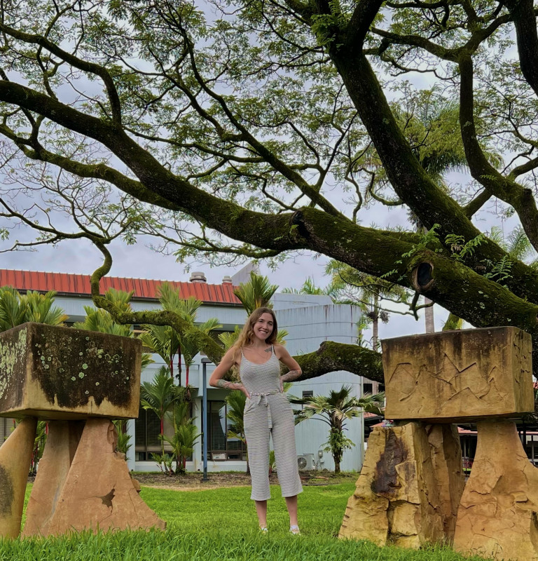 Darien standing and smiling between two rock structures. Behind her are palm trees and a campus building
